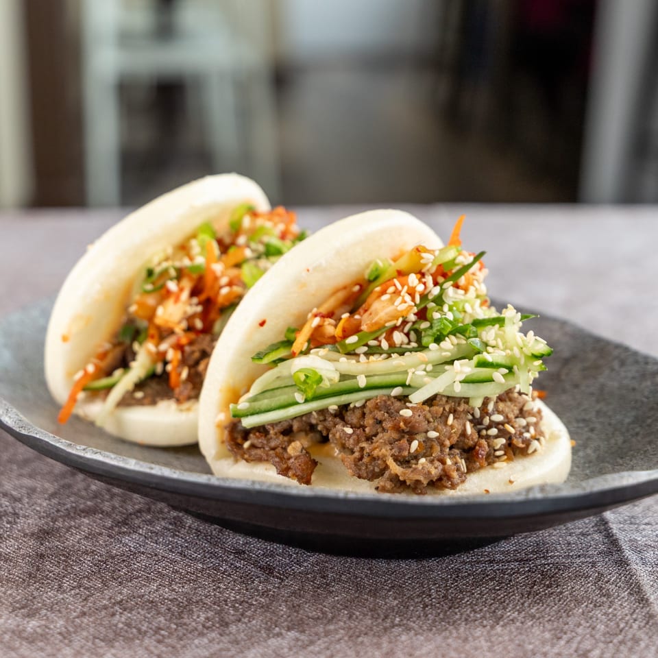 Two bao buns filled with ground meat, cucumber, kimchi, green onions, and sesame seeds.