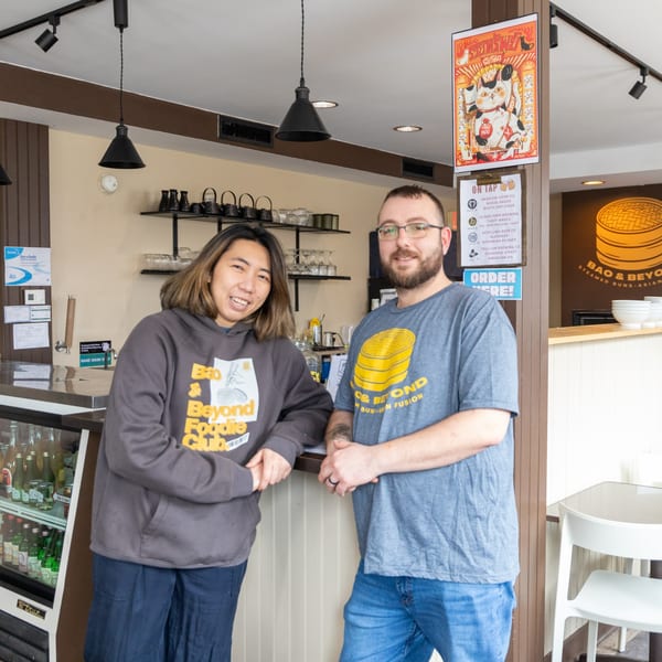 Two smiling people, a woman and a man, stand behind the counter of Bao & Beyond.