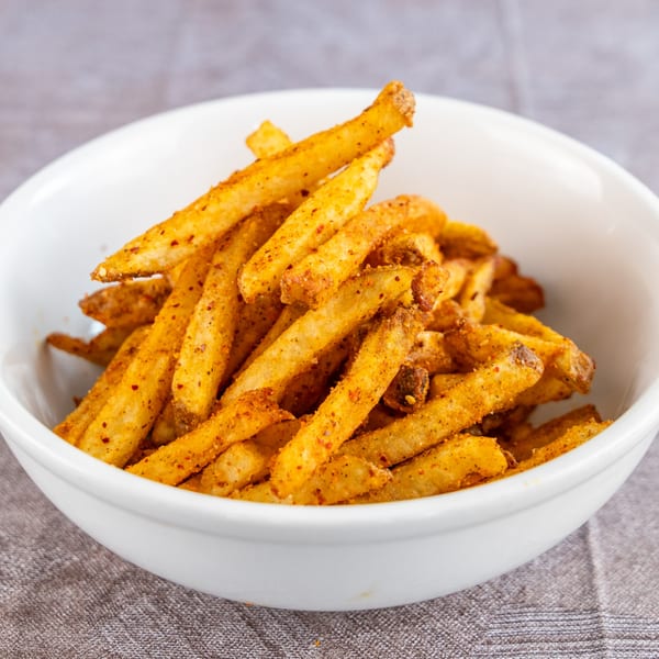 A white bowl filled with golden-brown seasoned french fries, resting on a gray tablecloth.