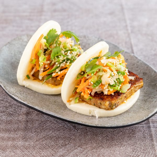 Two bao buns filled with crispy tofu, shredded carrots, cilantro, and sesame seeds on a gray plate.