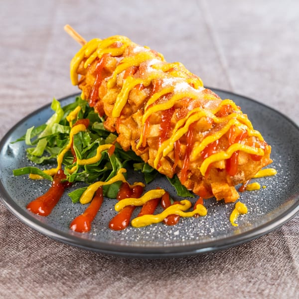 A Korean corn dog, topped with ketchup, mustard, and powdered sugar, rests on a bed of greens.