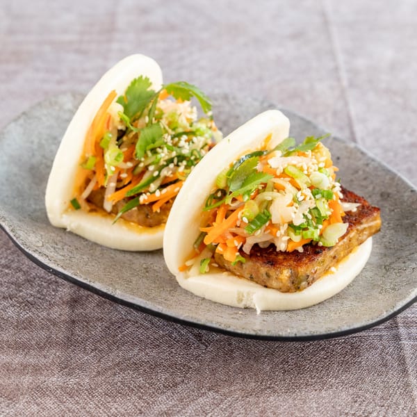 Two bao buns filled with grilled tofu, shredded carrots, green onions, cilantro, and sesame seeds.
