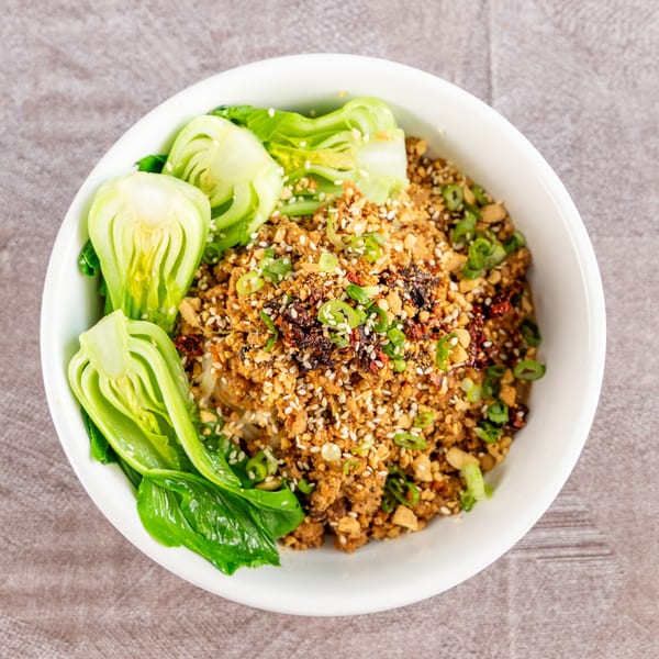 Overhead view of a white bowl filled with a savory dish, topped with peanuts, sesame seeds, and green onions, garnished with bok choy.