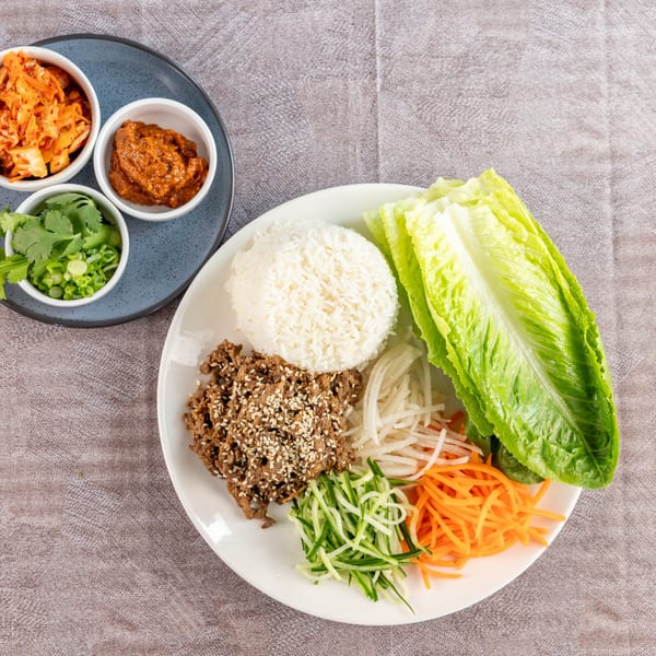 Korean BBQ lettuce wraps with bulgogi, rice, vegetables, kimchi, and sauces on a gray tablecloth.