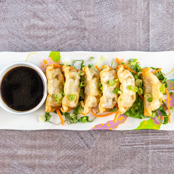 Fried dumplings with green onions and shredded carrots on a white plate with a side of soy sauce.