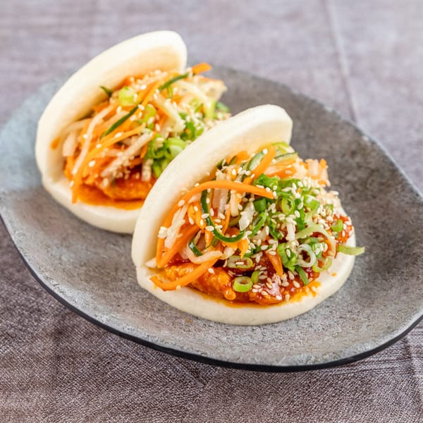 Two bao buns filled with chicken, shredded carrots, cucumbers, and green onions, topped with sesame seeds.
