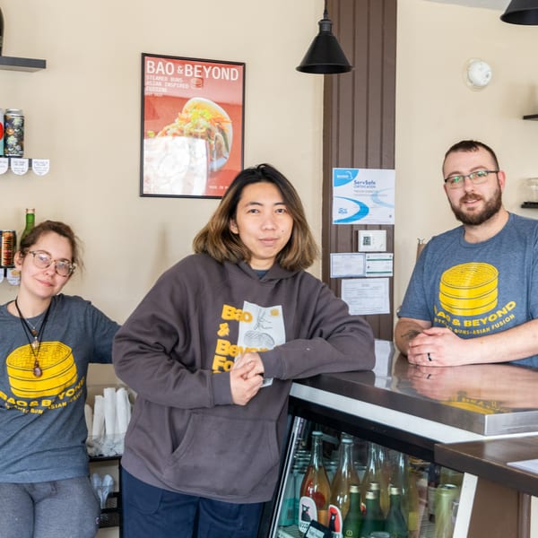 Three Bao & Beyond staff members, two women and one man, stand behind the counter.