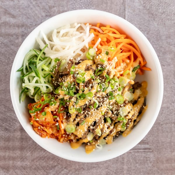 A white bowl filled with bulgogi, kimchi, shredded carrots, cucumbers, and daikon radish, drizzled with sauce.