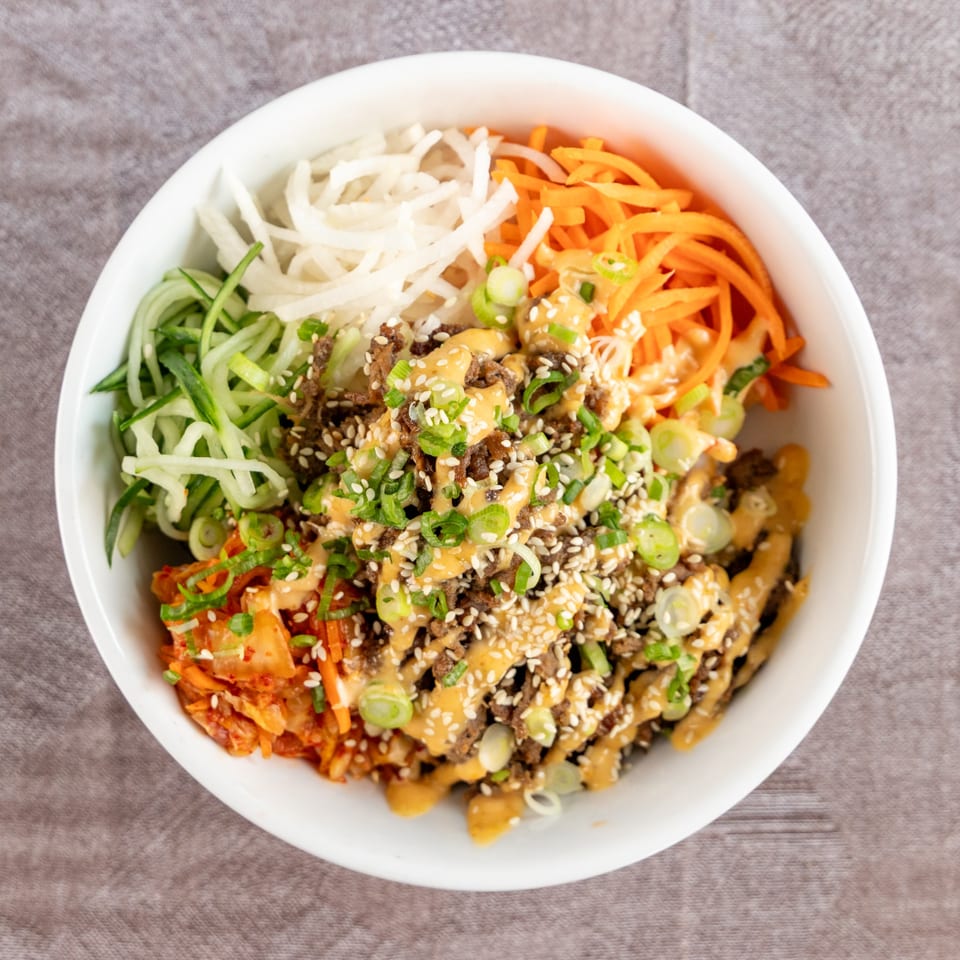A white bowl filled with bulgogi, kimchi, shredded carrots, cucumbers, and daikon radish, drizzled with sauce.