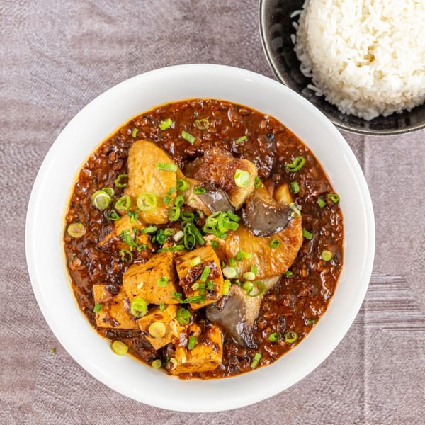A white bowl of braised tofu and mushrooms in a dark sauce, garnished with green onions, next to white rice.