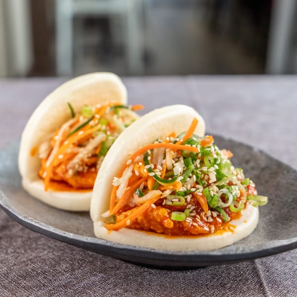 Two bao buns filled with orange chicken, shredded carrots, cabbage, green onions, and sesame seeds.