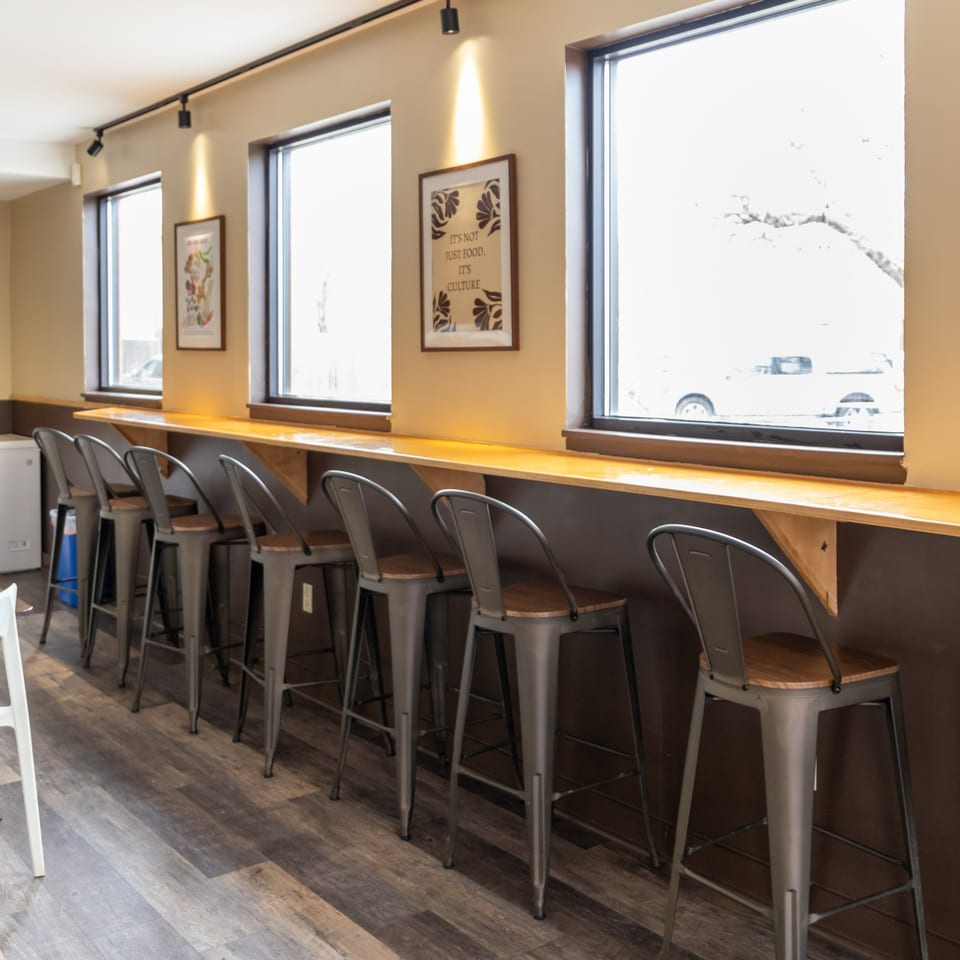 A row of metal bar stools with wooden seats lines a window counter in a cafe.